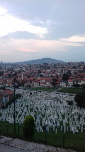 A picture of the city taken from a hilltop. In the foreground, a crematorium with many white gravestones.